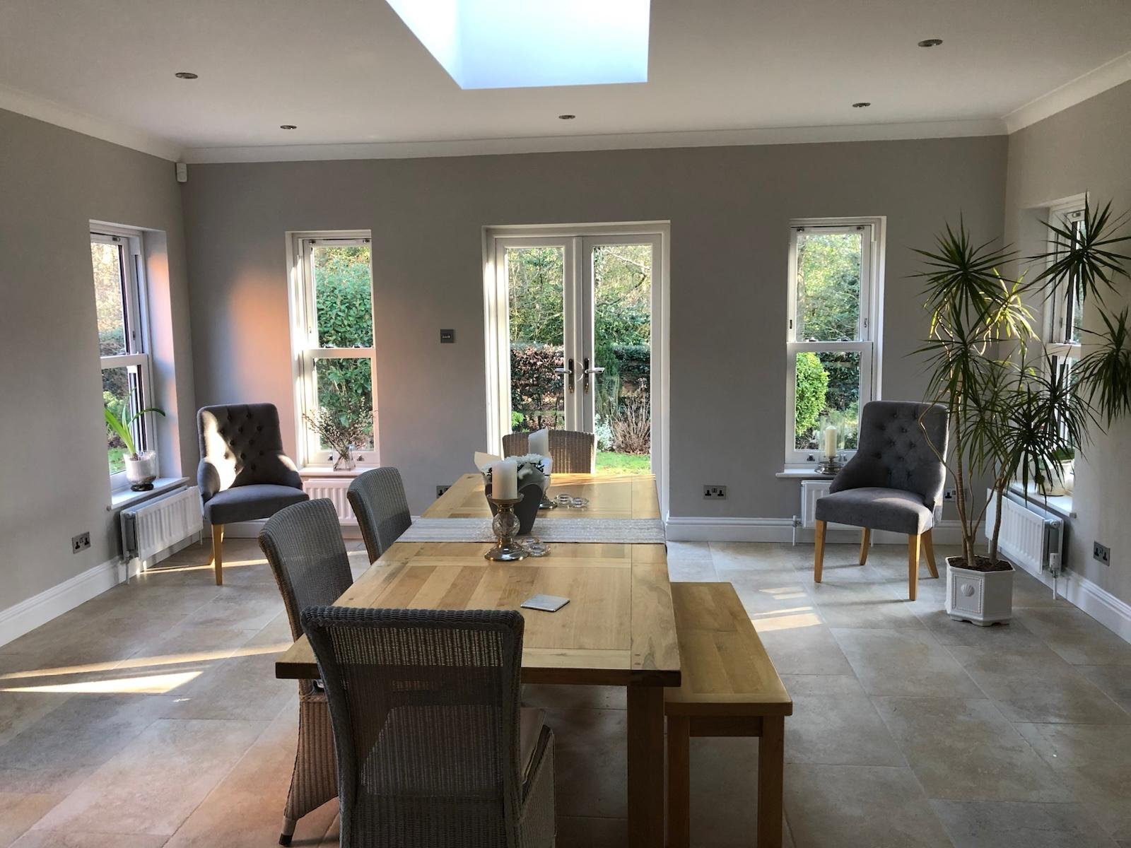 Modern dining room with wooden table, black chairs, large windows, skylight, and potted plants overlooking garden