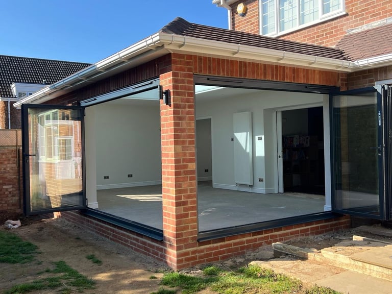 Modern glass-fronted conservatory extension attached to brick house with grey roof and large sliding doors