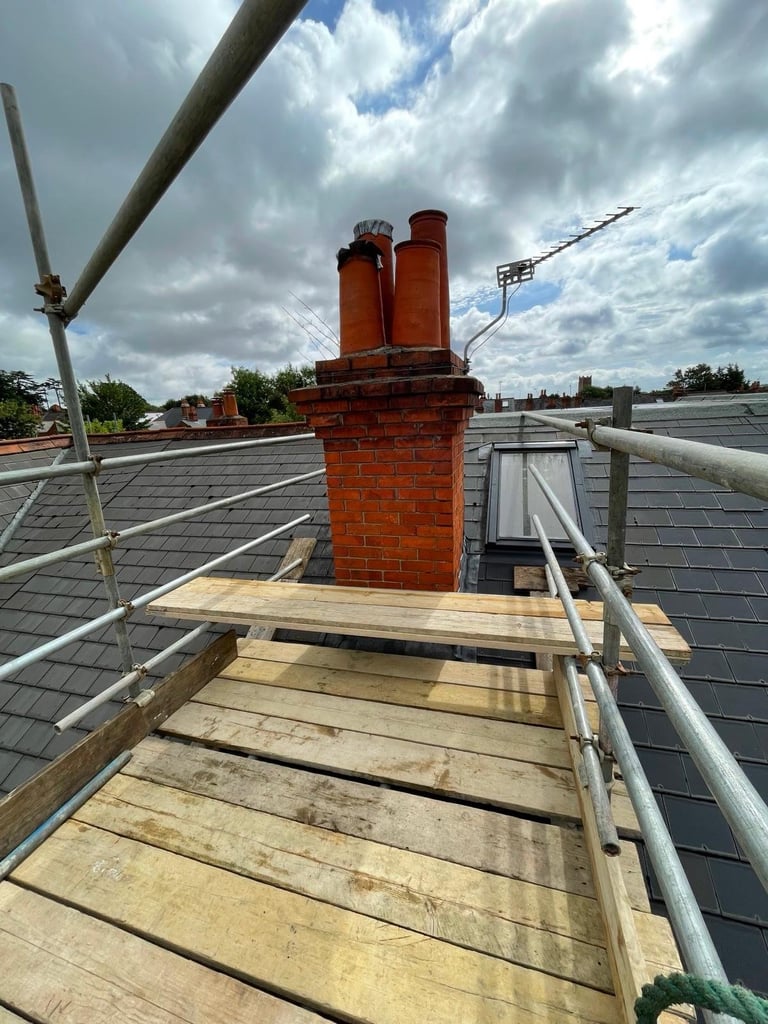 Red brick chimney with metal vent pipes on residential roof with wooden walkway and metal railing under cloudy sky