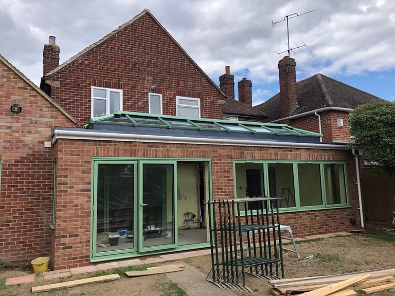 Brick house with green-framed glass extension and metal railing under construction on a cloudy day