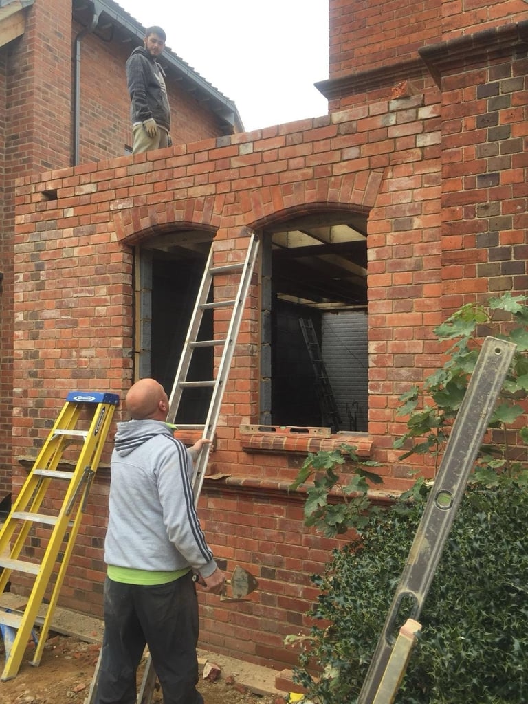 Construction workers on ladders working on brick building with two arched window openings and ivy growing on the right side