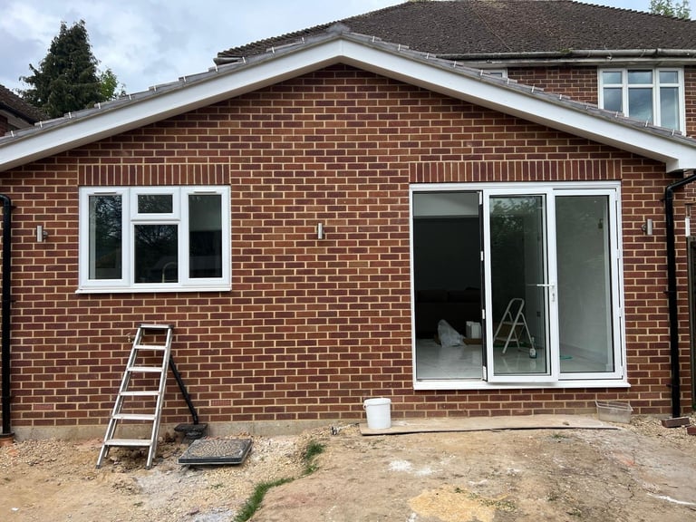 New brick home extension with white-framed windows and sliding glass doors under construction with ladder and materials on ground