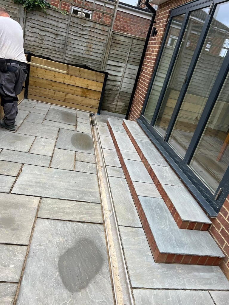 Patio during installation with gray flagstone pavers, red brick edging, and large modern black-framed doors against a brick wall