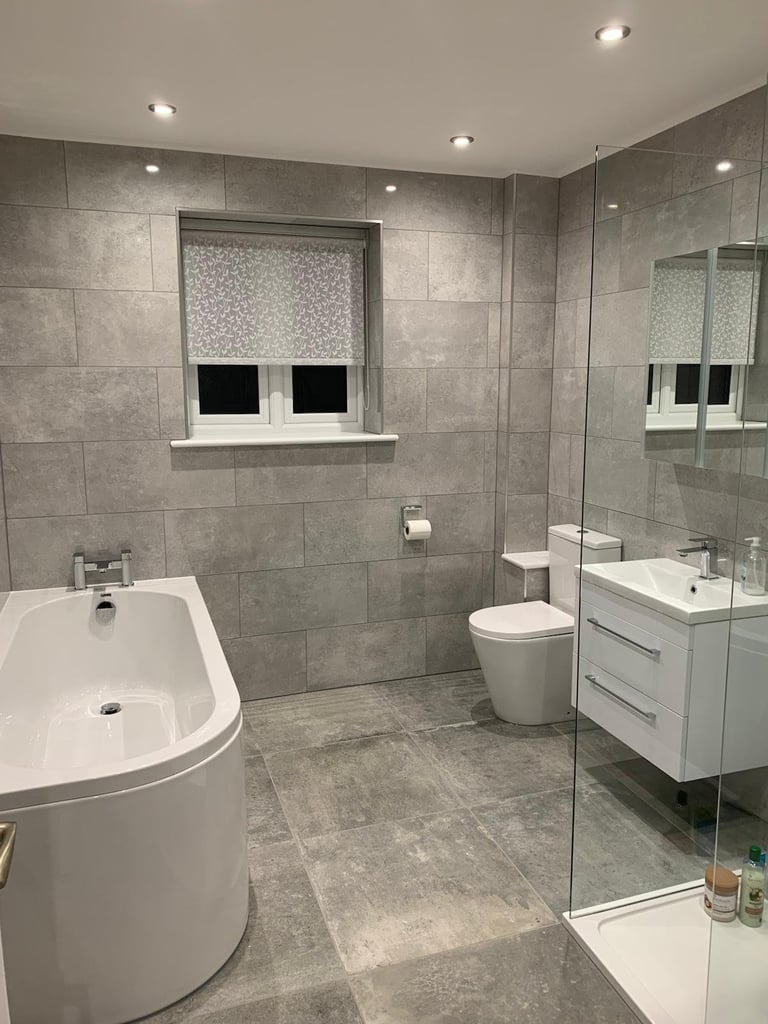 Modern bathroom with gray tile walls, white curved soaking tub, white vanity, toilet, and glass shower enclosure with window