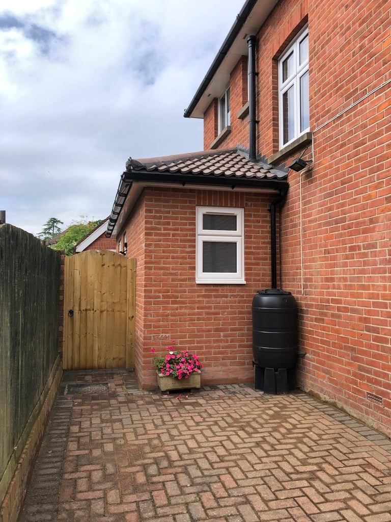 Red brick residential extension with white window, patterned roof, wooden gate, flowering planter, and water butt beside paved courtyard